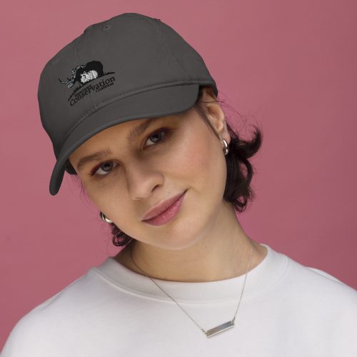 VCC: Woman wearing an organic baseball hat with "conservation" text and rhino graphic, looking at the camera against a pink background.