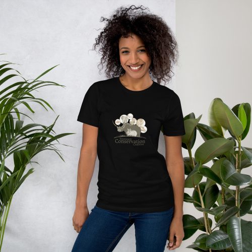 VCC: Sentence with product name: A smiling woman with curly hair wearing a black Earthly unisex t-shirt with a "conservation" print, standing in front of a potted plant background.